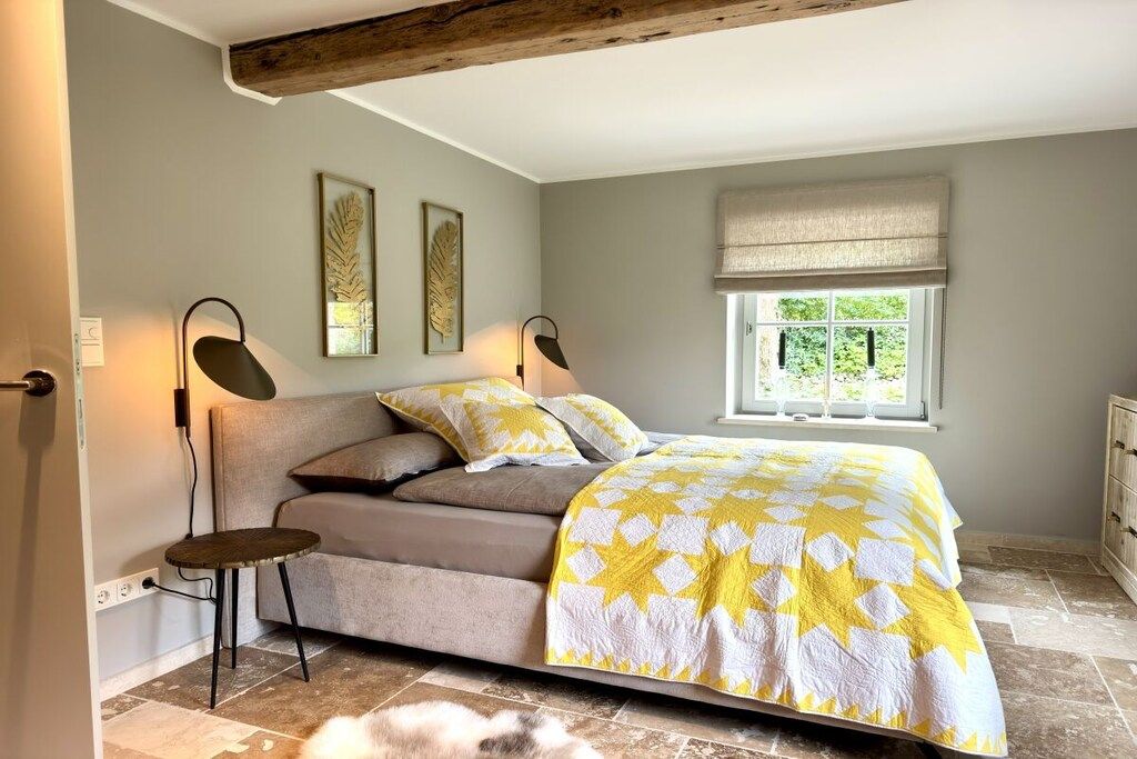 Ein Schlafzimmer mit einem Bett, einem Nachttisch, einer Lampe und einem Fenster mit Blick ins Grüne.