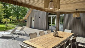 Een terras met een houten tafel en stoelen, een hanglamp en uitzicht op een gebouw met verticale beplating.