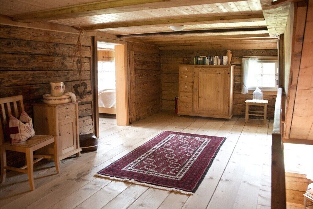 Ein Holzblockhaus-Innenraum mit Holzboden, einem roten Teppich mit Muster, einem Holzschrank und einem Fenster mit Vorhängen.