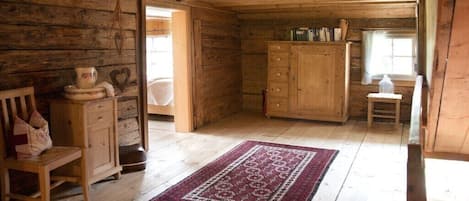 A wooden cabin interior with a wooden floor, a red patterned rug, a wooden cabinet, and a window with curtains.
