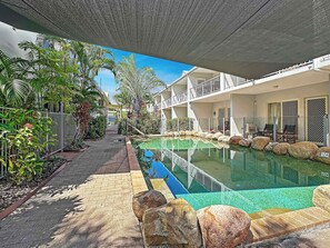 A pool area with a clear view of the pool, surrounding plants, and a building with balconies. - Sails On Horseshoe Apartment 10 (Horseshoe Bay)