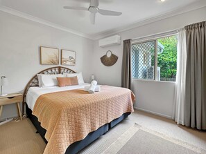 A bedroom with a bed, a bedside table, a ceiling fan, an air conditioning unit, a window with curtains, and artwork on the wall. - Sails On Horseshoe Apartment 10 (Horseshoe Bay)