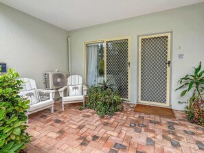 Un patio couvert avec une chaise blanche, une petite table et une plante en pot. Il y a deux portes équipées de moustiquaires et une enseigne sur le mur.