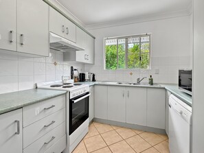 A modern kitchen with white cabinetry, a built-in oven, a stovetop, and a window with a view of greenery. - Sails On Horseshoe Apartment 10 (Horseshoe Bay)