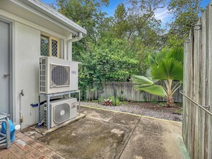 A residential backyard with a white house, outdoor air conditioning units, a wooden fence, and a palm tree. - Sails On Horseshoe Apartment 10 (Horseshoe Bay)