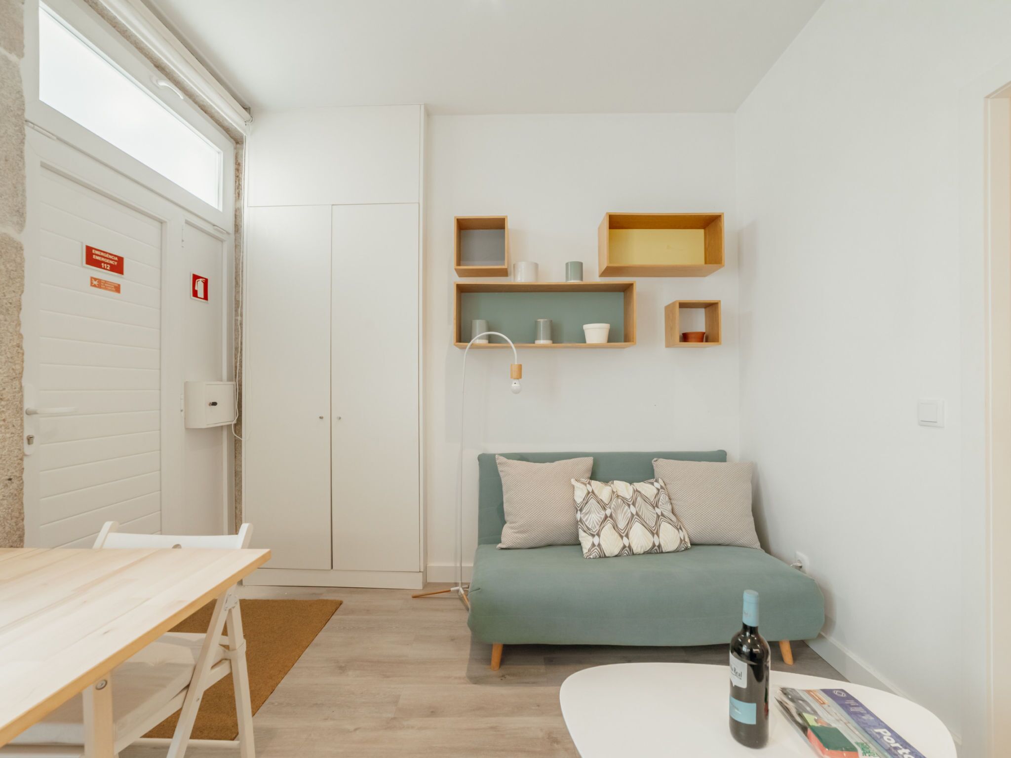 A small, well-lit room with a wooden table, a green sofa with patterned cushions, and a wine bottle on a small table. There are shelves with decorative items and a door with emergency signage.