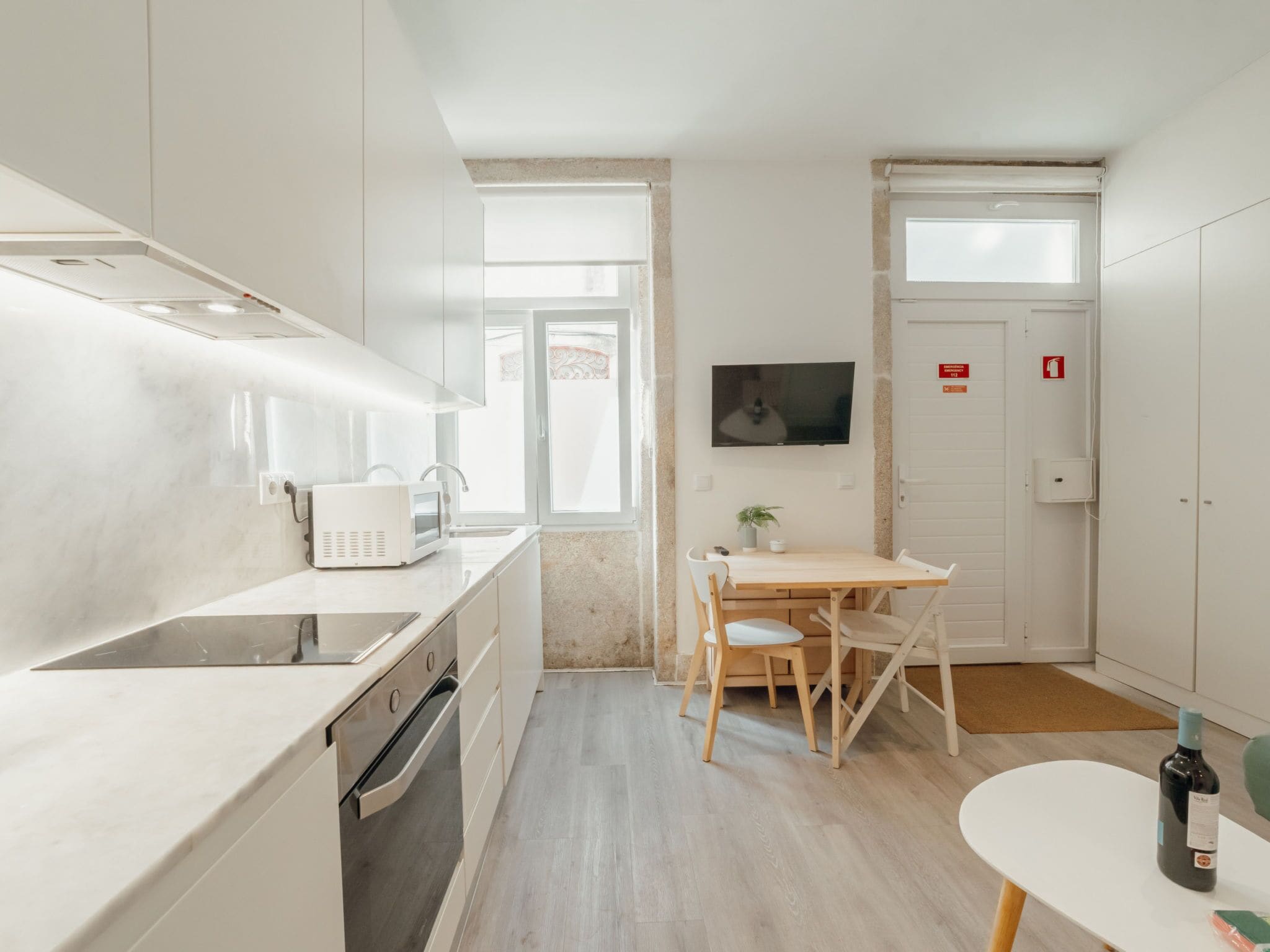 Eine kompakte Küche mit Backofen, Mikrowelle und Kaffeemaschine. Es gibt eine kleine Essecke mit Tisch und Stühlen, und ein an der Wand befestigtes Fernsehgerät.