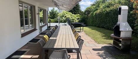 Un patio al aire libre con una mesa de madera larga y sillas, una parrilla y un área de estar techada.