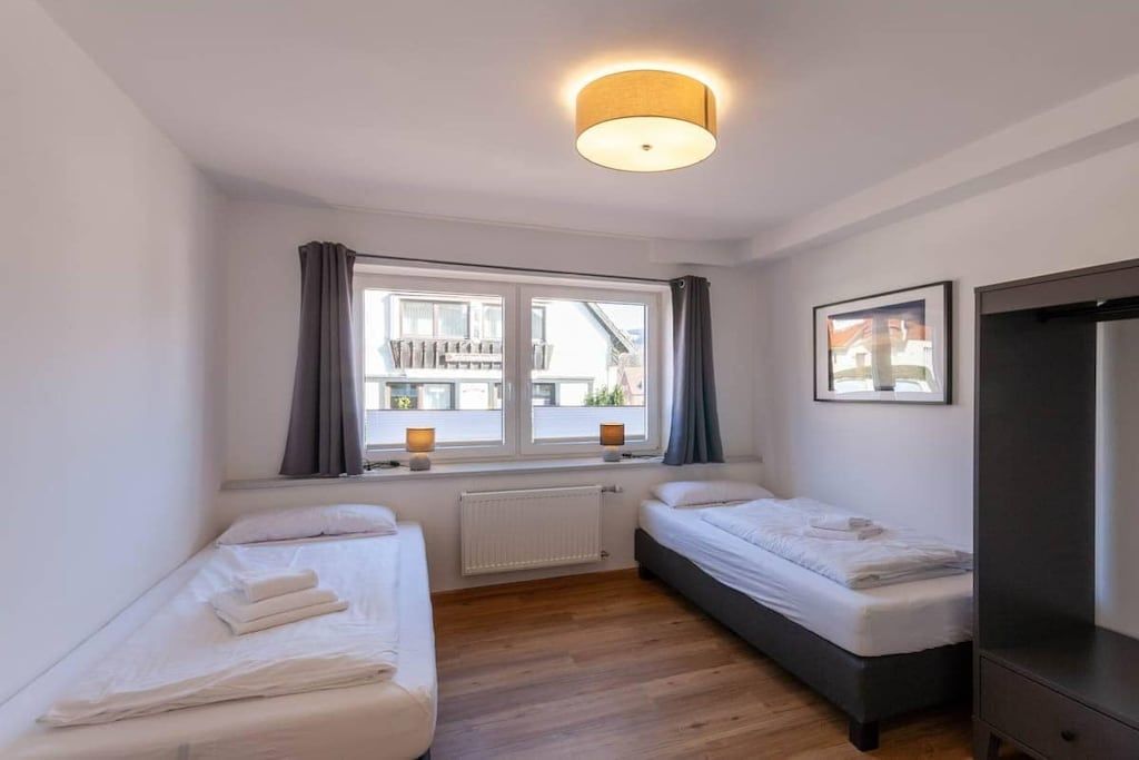 Habitació petita i neta amb dos llits individuals, una finestra amb cortines, un radiador i un armari.