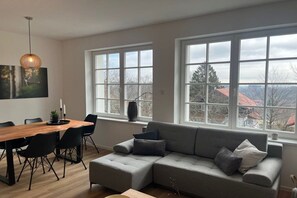 A modern living room with a grey sectional sofa, a wooden dining table with chairs, and large windows offering a view of the outside. - Holiday apartment with 3 bedrooms (Braunlage)
