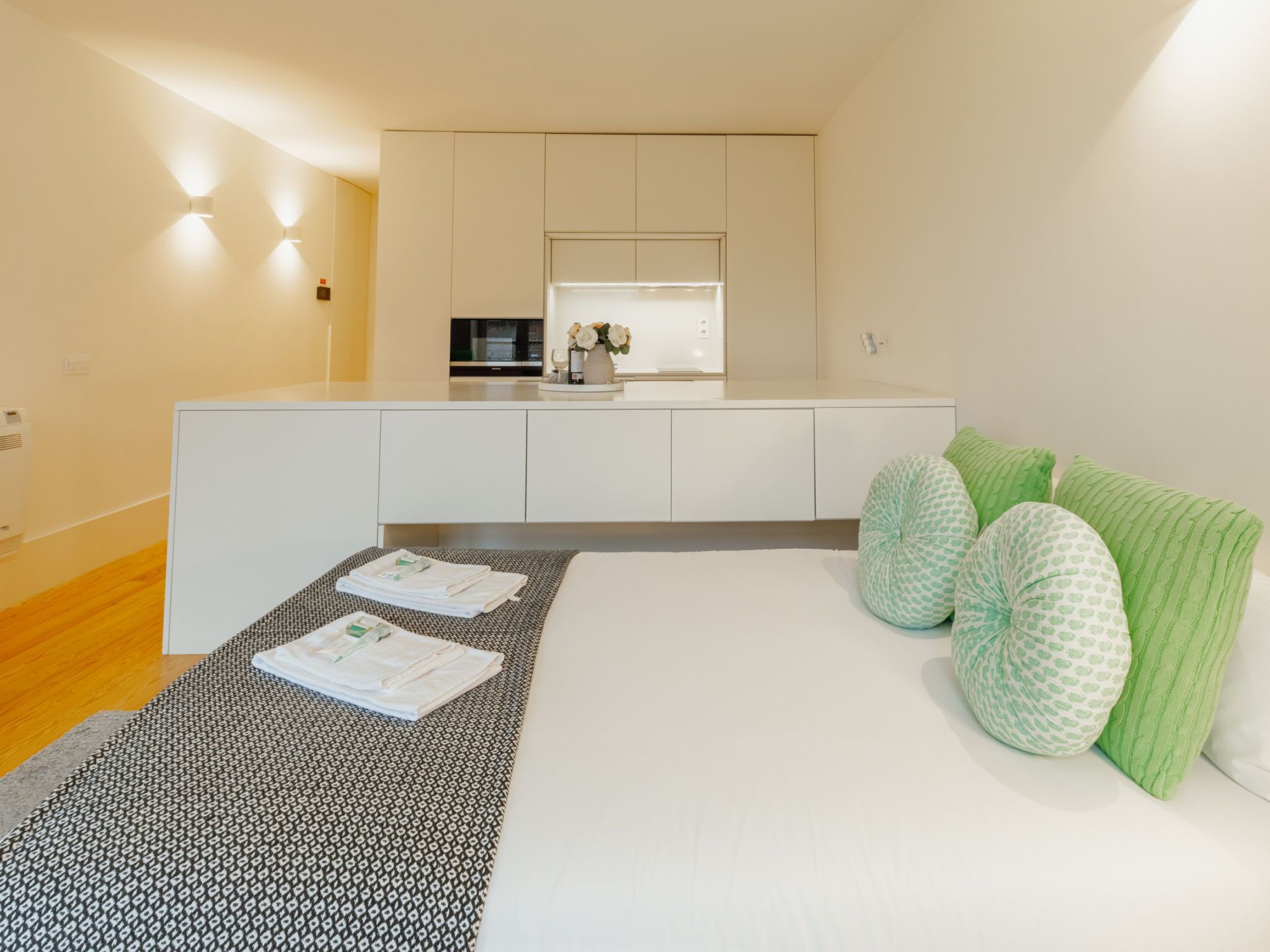 A modern bedroom with a bed, pillows, a bedside table, and a small kitchenette area.