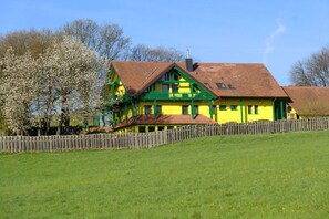 Une maison à deux étages, avec une façade verte et jaune, une clôture en bois et une cour couverte d’herbe.