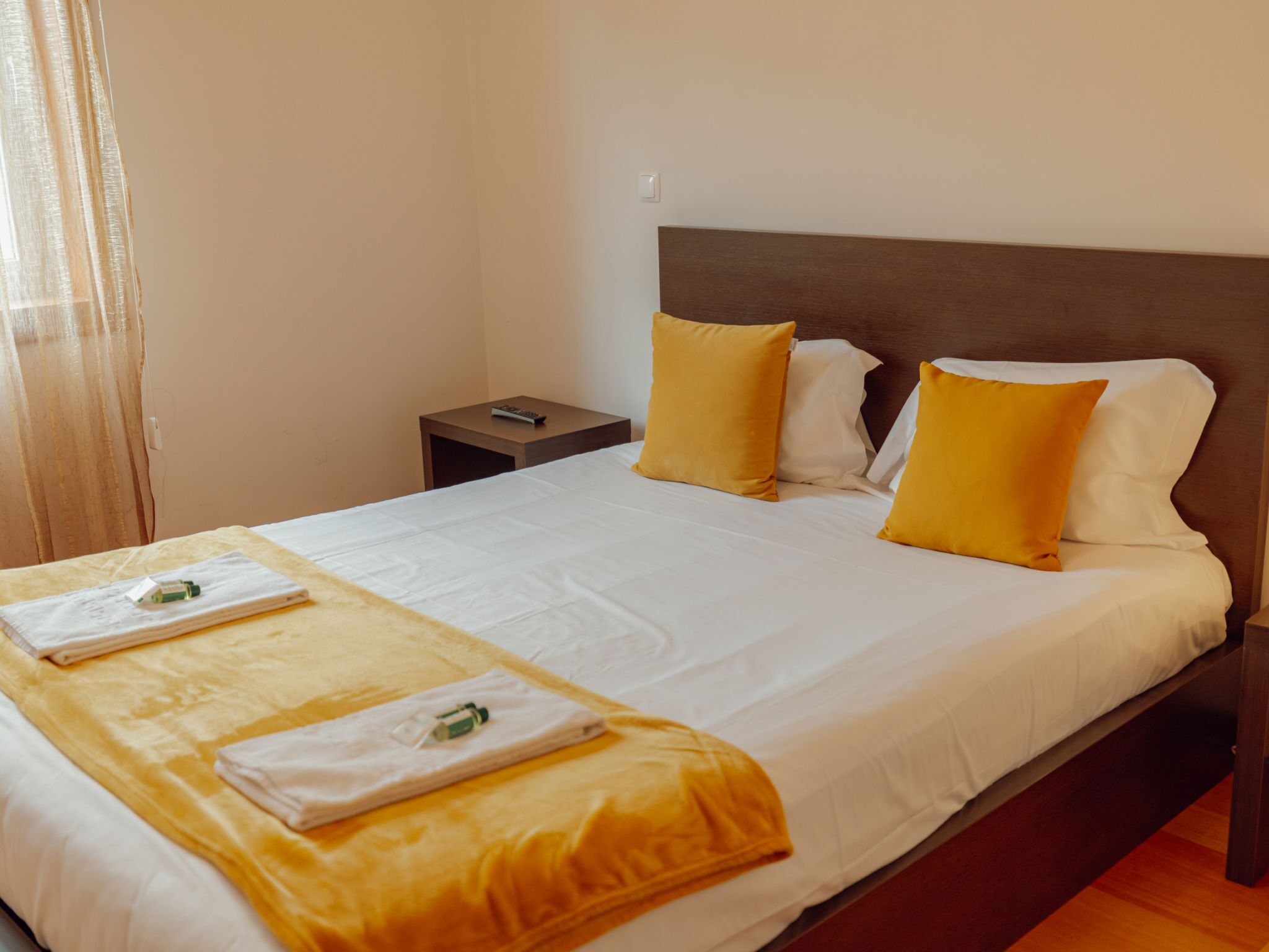 A neatly made bed with white and yellow linens, a wooden headboard, and a small bedside table with a remote control.