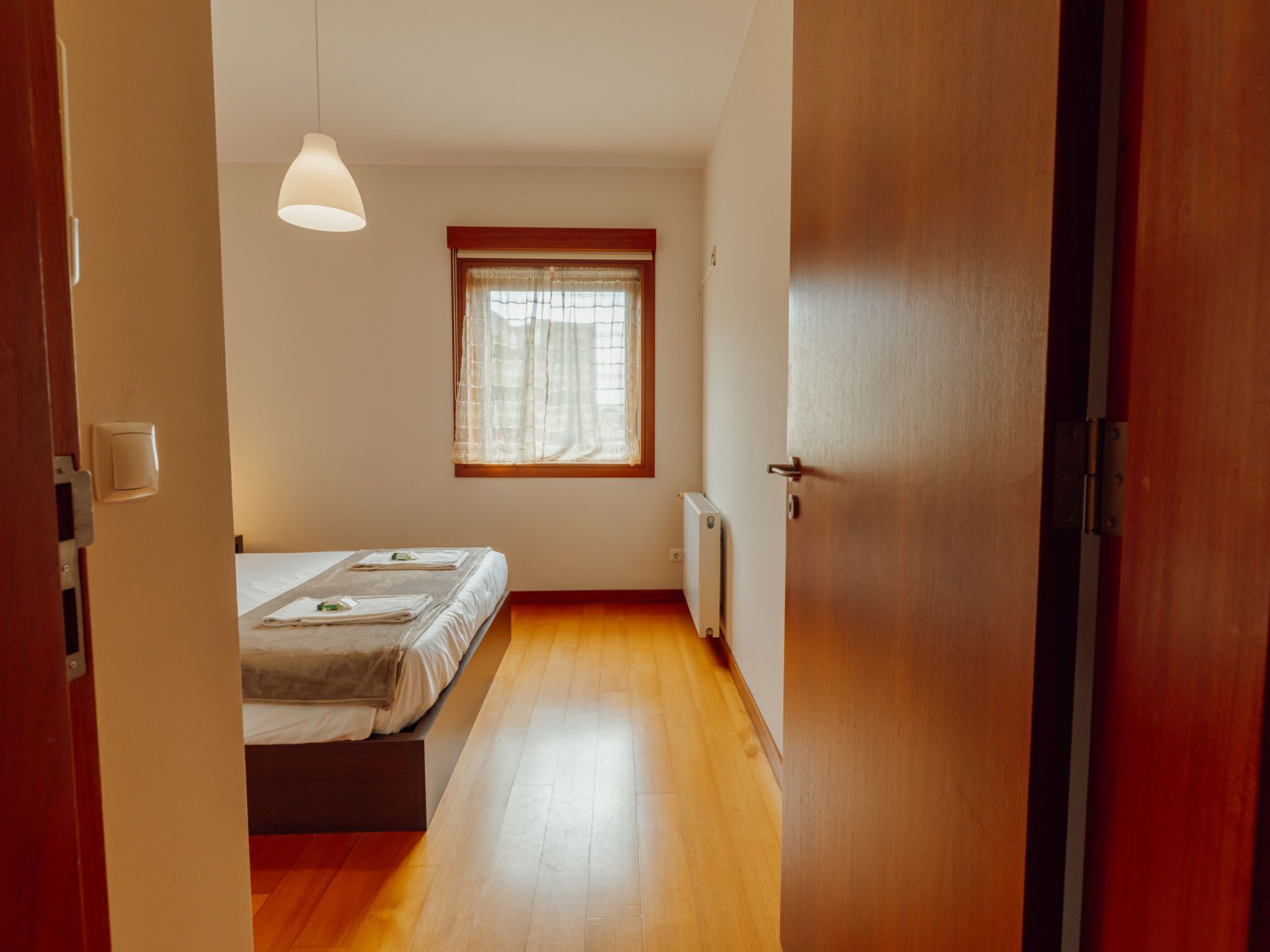 Un pasillo estrecho que da a un dormitorio con cama, una ventana con cortinas, un radiador y una puerta.