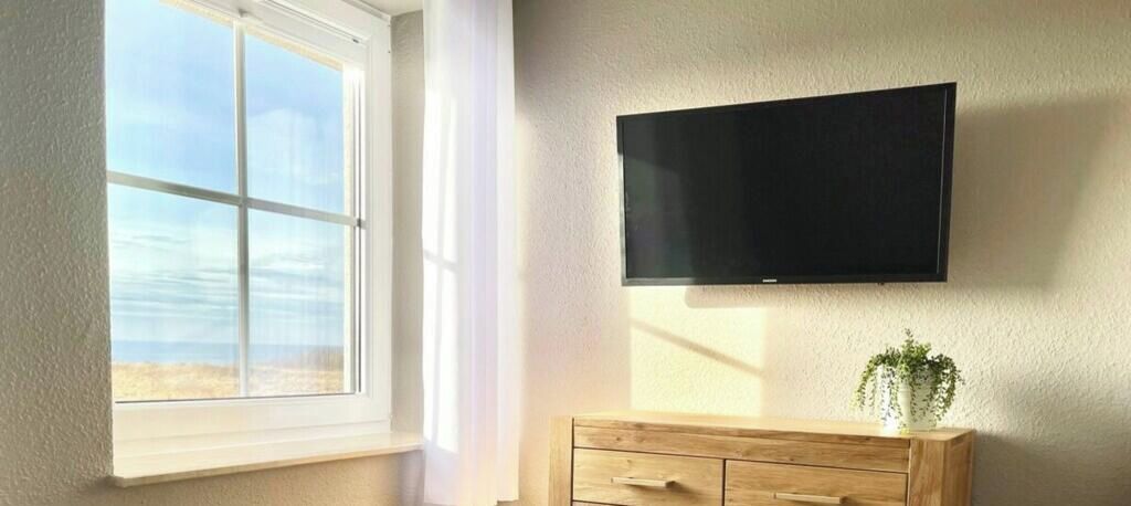 A room with a large window, a flat-screen TV mounted on the wall, and a wooden cabinet with drawers.