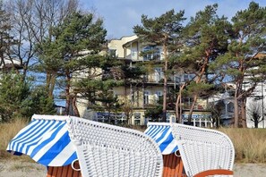 Blick auf einen Strand mit einem Gebäude im Hintergrund, zwei Korbstühle mit gestreiften Kissen und Kiefern.