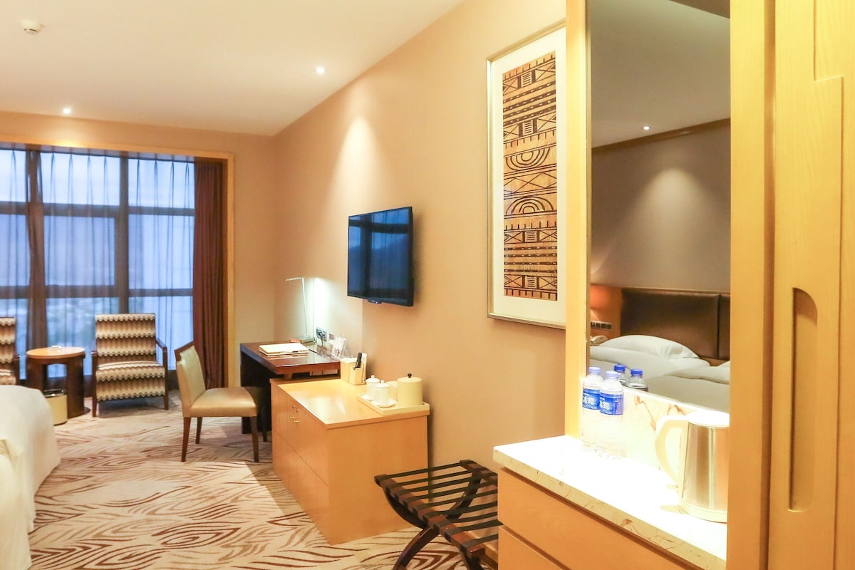 A hotel room with a desk, chair, bed, and a large window.