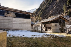 A rustic cabin with a sloped roof, surrounded by snow-covered ground and a mountainous backdrop.