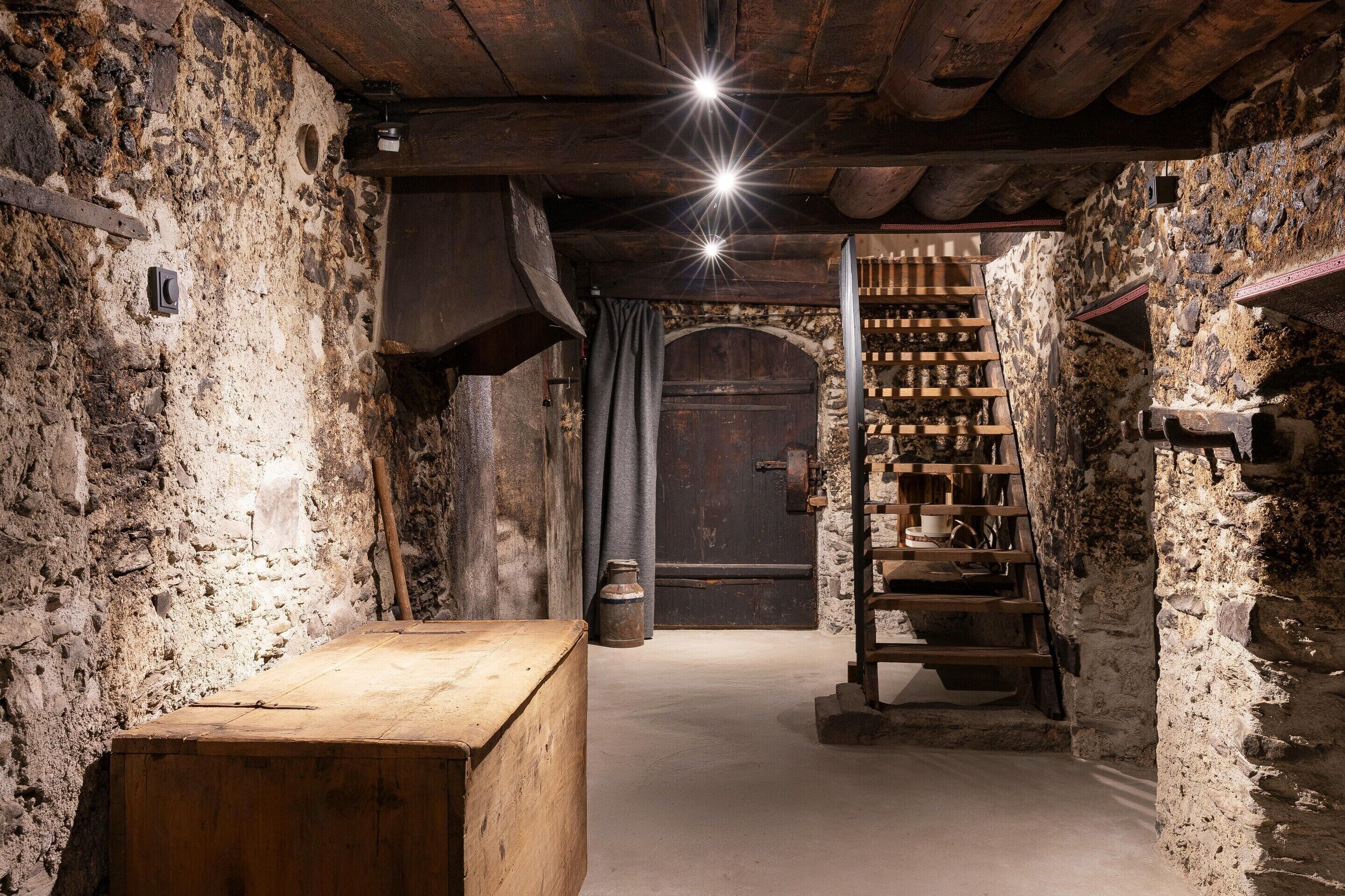 A narrow stone corridor with a wooden bench, a wooden ladder, and a dark wooden door at the end.