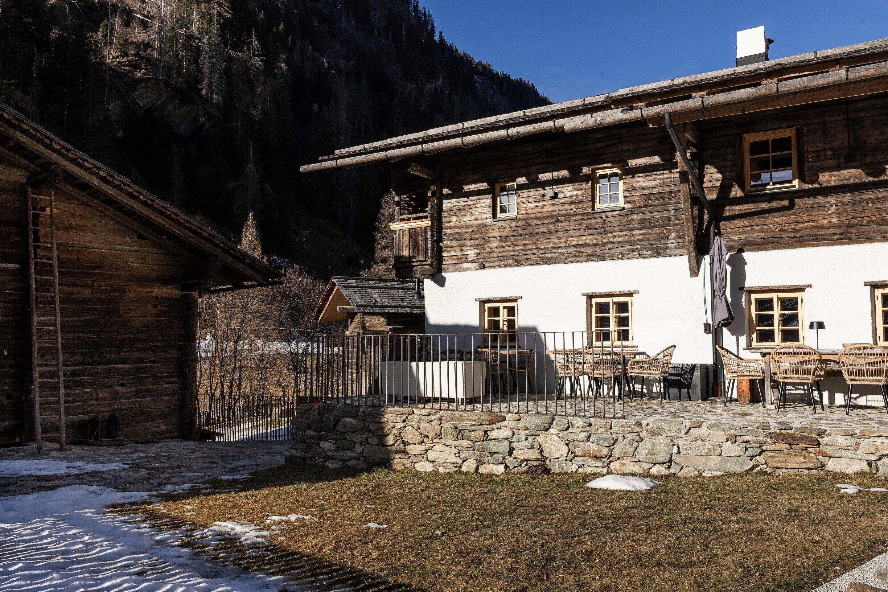 A rustic wooden building with a stone wall, outdoor seating, and a mountain backdrop.