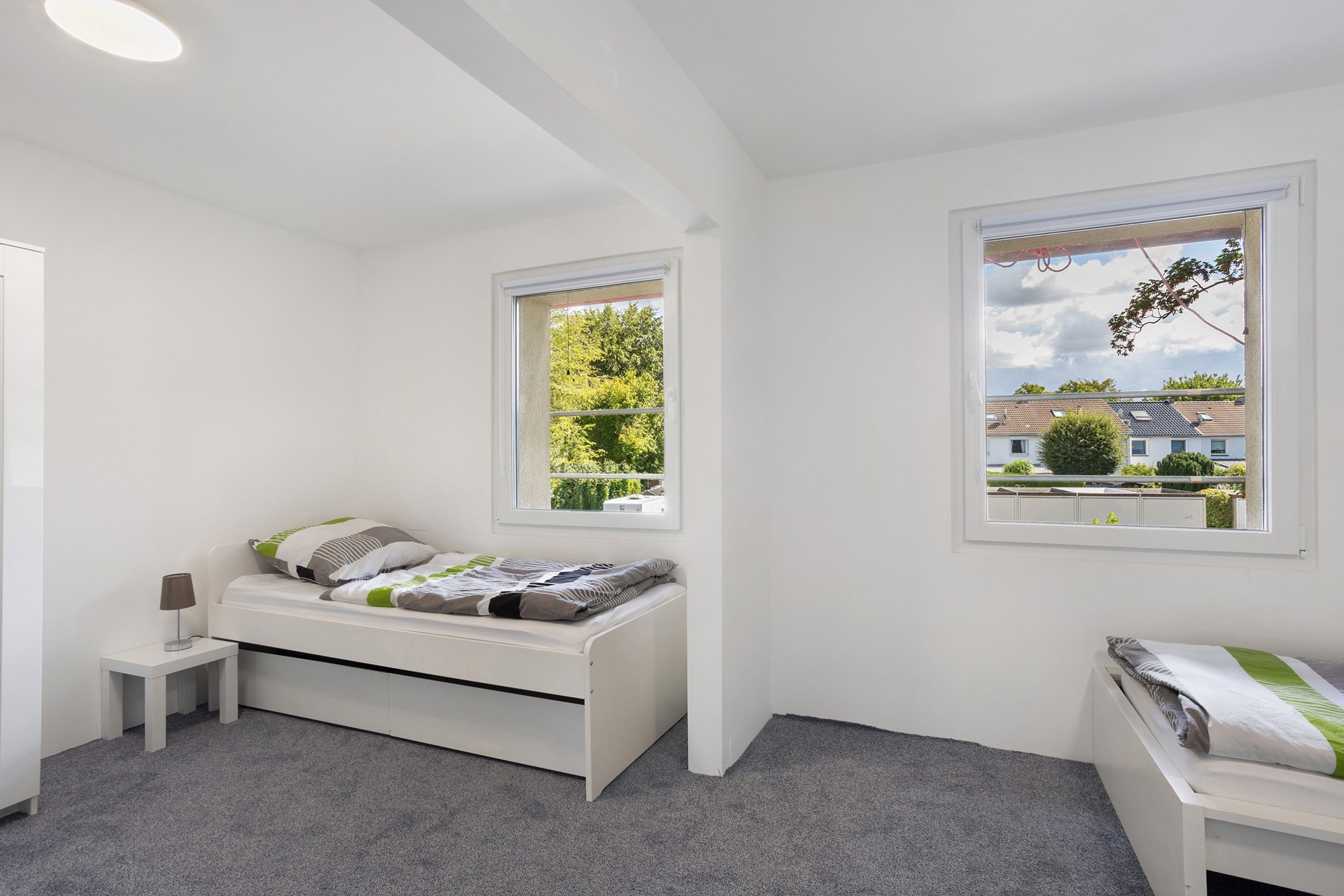 A room with a bed, a nightstand, a lamp, and two windows with views of greenery and buildings.