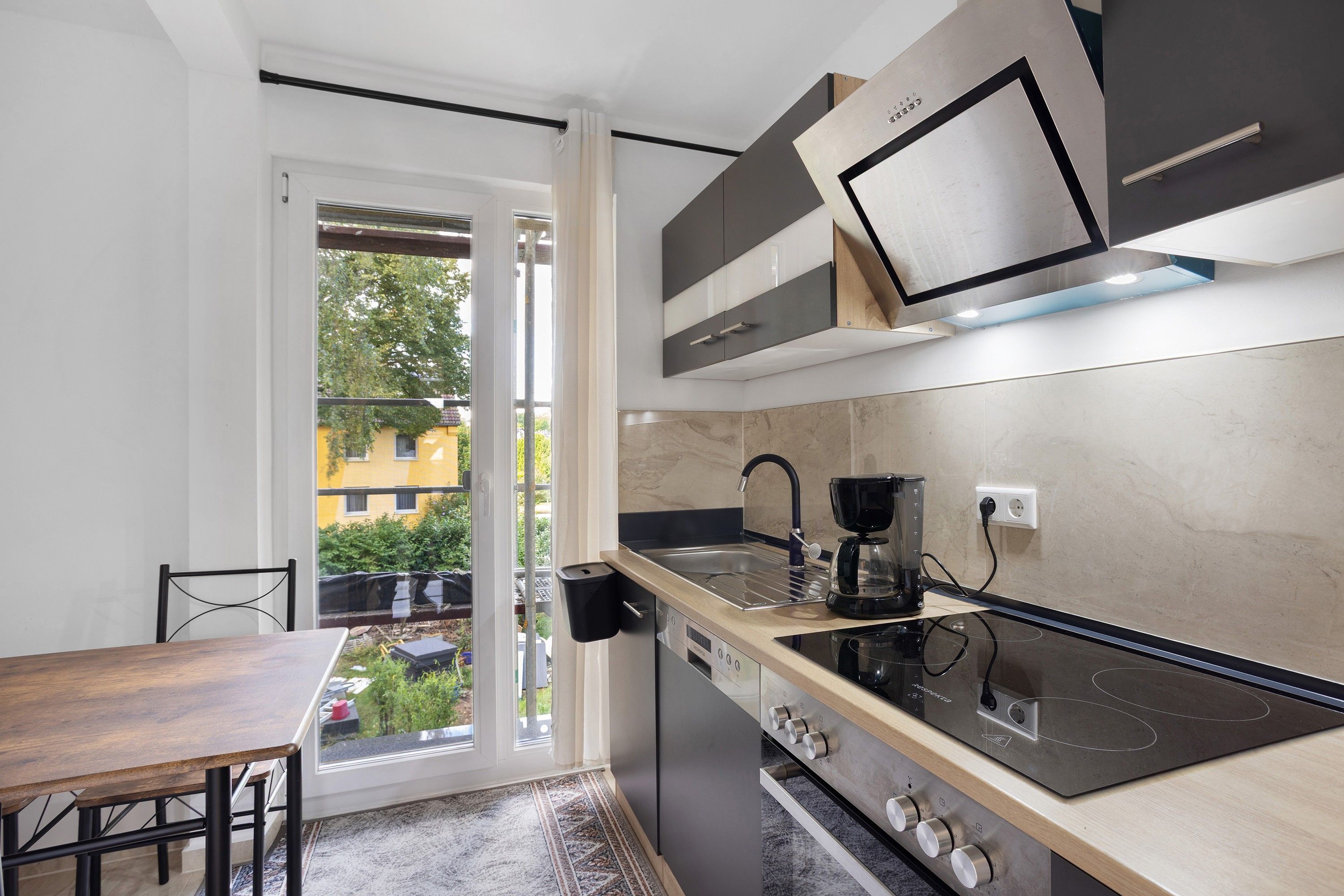 Une cuisine moderne équipée d’une cuisinière intégrée, d’une machine à café et offrant une vue sur un immeuble jaune à travers la porte vitrée.