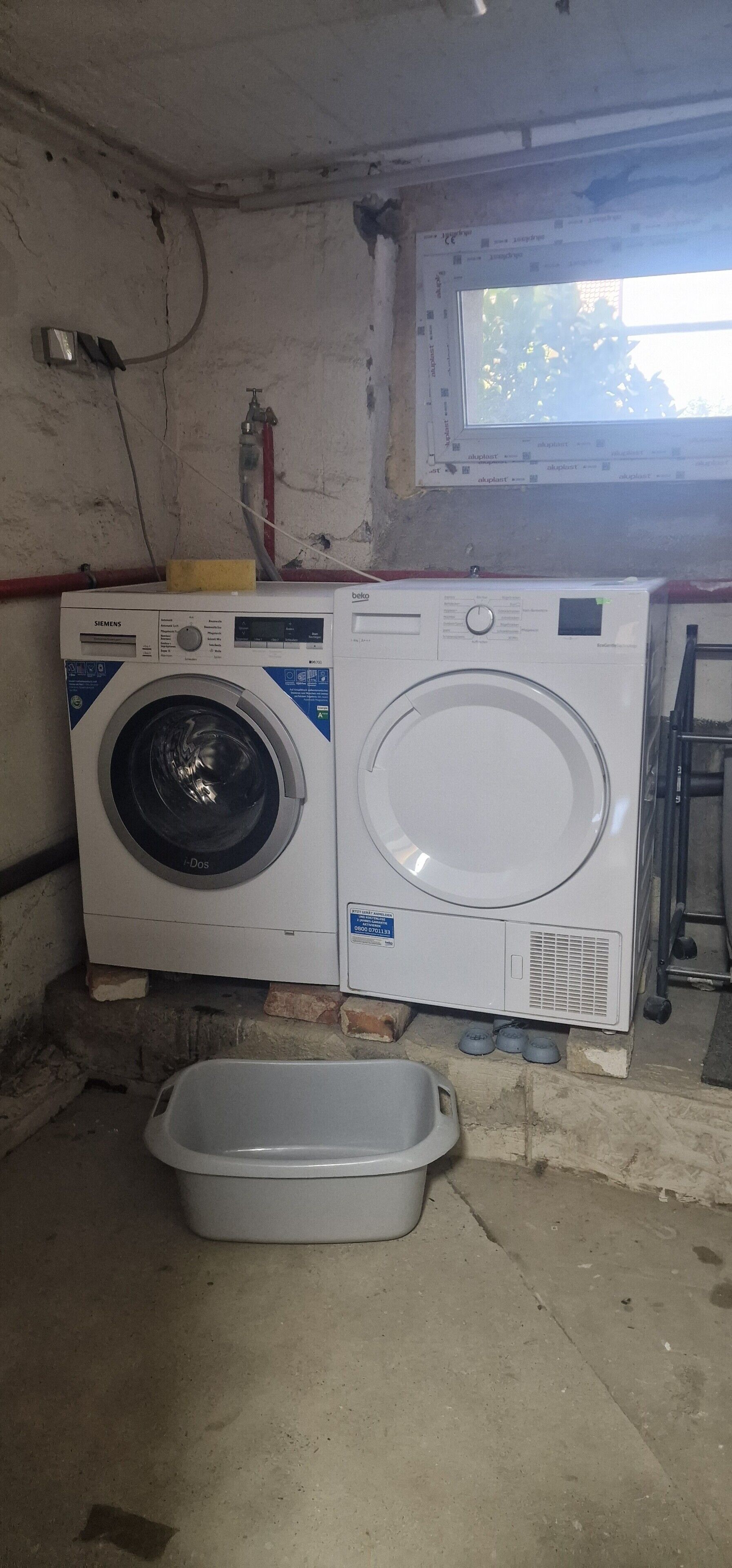A washing machine and a plastic basin in a room with a window and a ladder.