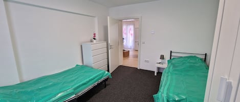 Une petite pièce étroite, avec deux lits simples recouverts de draps verts, une commode blanche et une petite table avec une lampe.