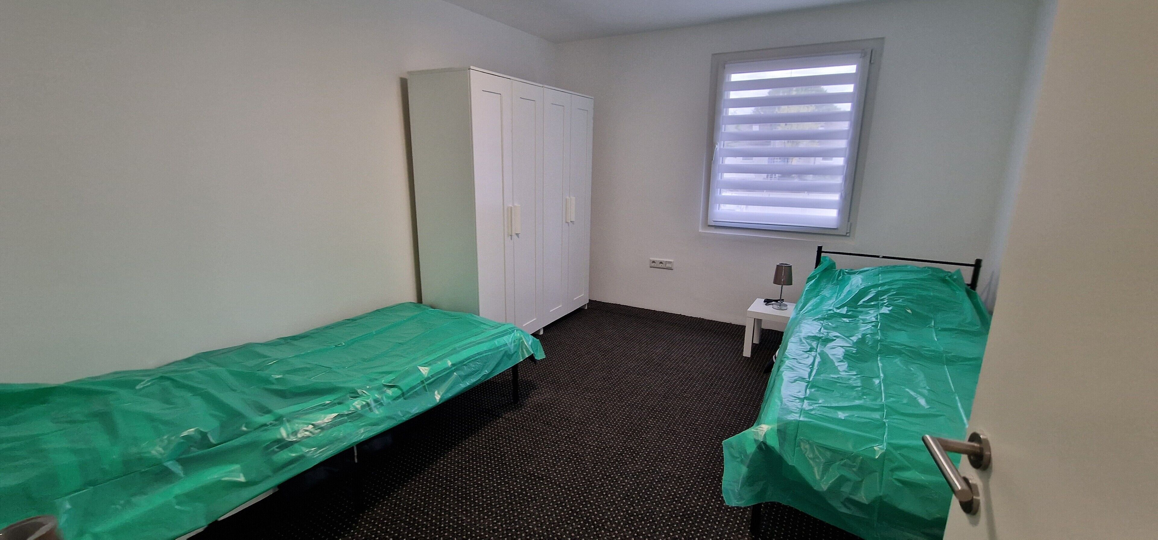 A small, single-room accommodation with two beds, a wardrobe, a window with blinds, and a small table with a lamp.