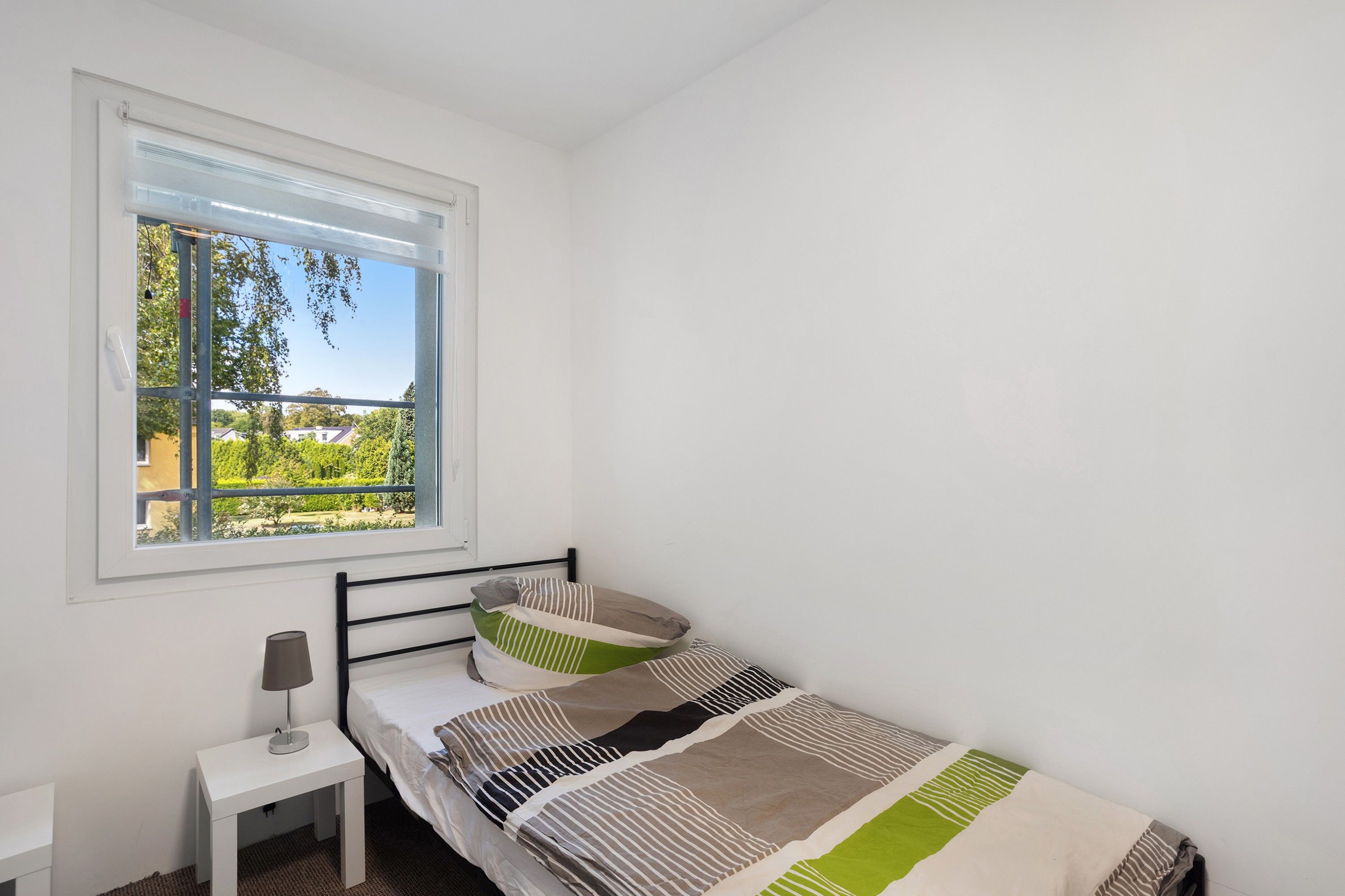 Un dormitorio con cama, mesita de noche con lámpara y ventana con vistas a zonas verdes y edificios.