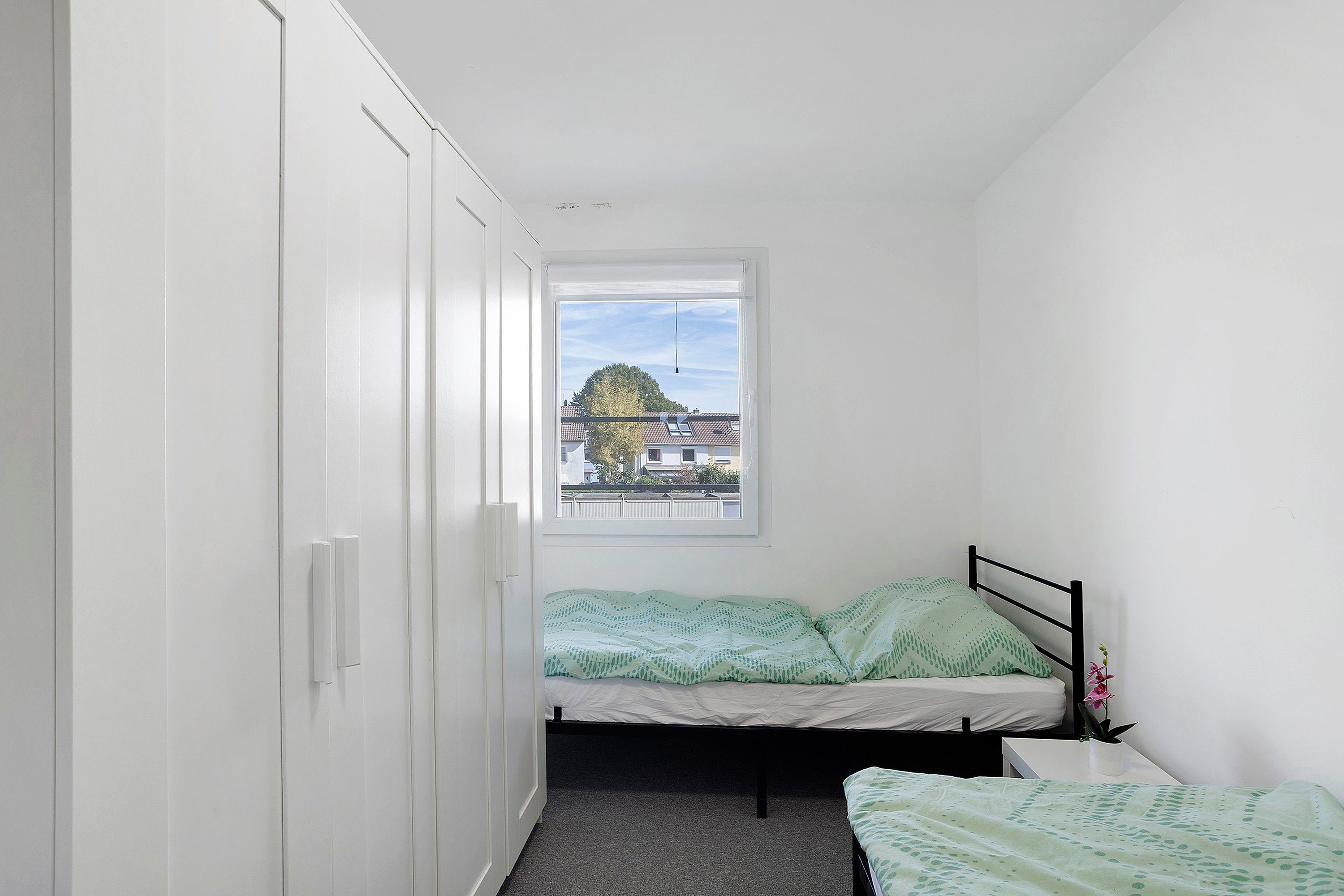 A small, narrow room with a single bed, a window, and a wardrobe.