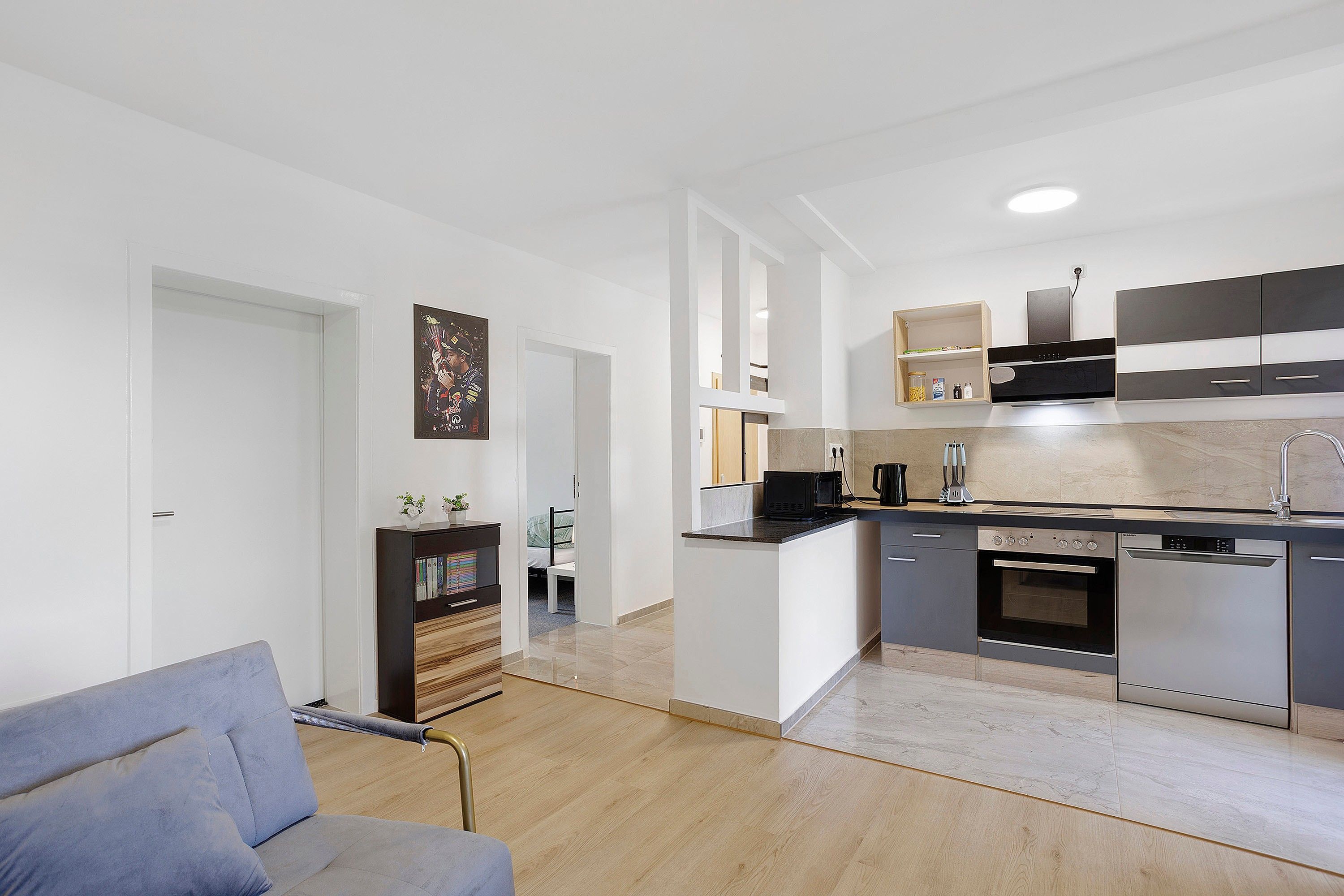 A modern kitchen with a built-in oven, microwave, and a coffee machine. There is a dining area with a grey sofa and a bookshelf.