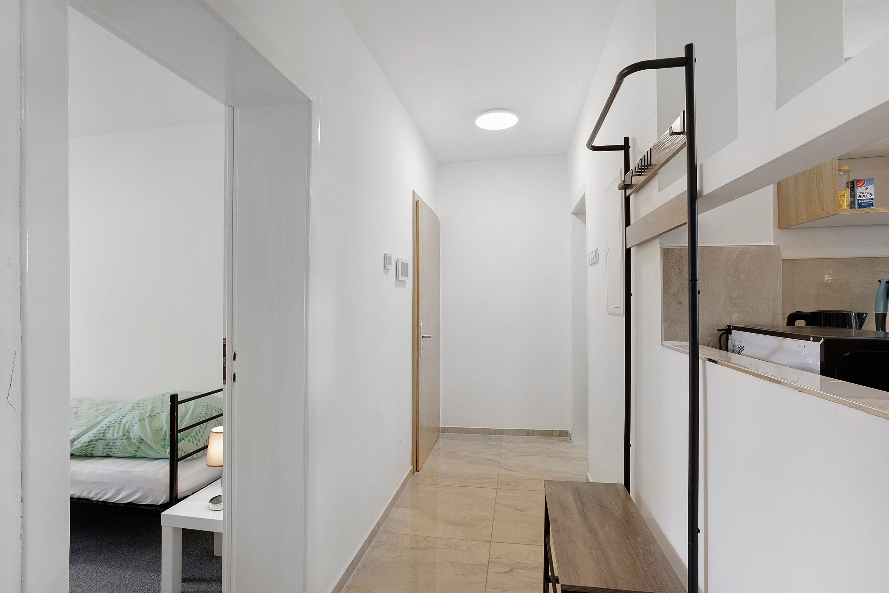 A narrow hallway with a bed, a small table, and a bench. There is a kitchenette area with a sink and a stove.