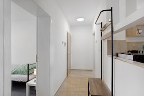 A narrow hallway with a bed, a small table, and a bench. There is a kitchenette area with a sink and a stove.
