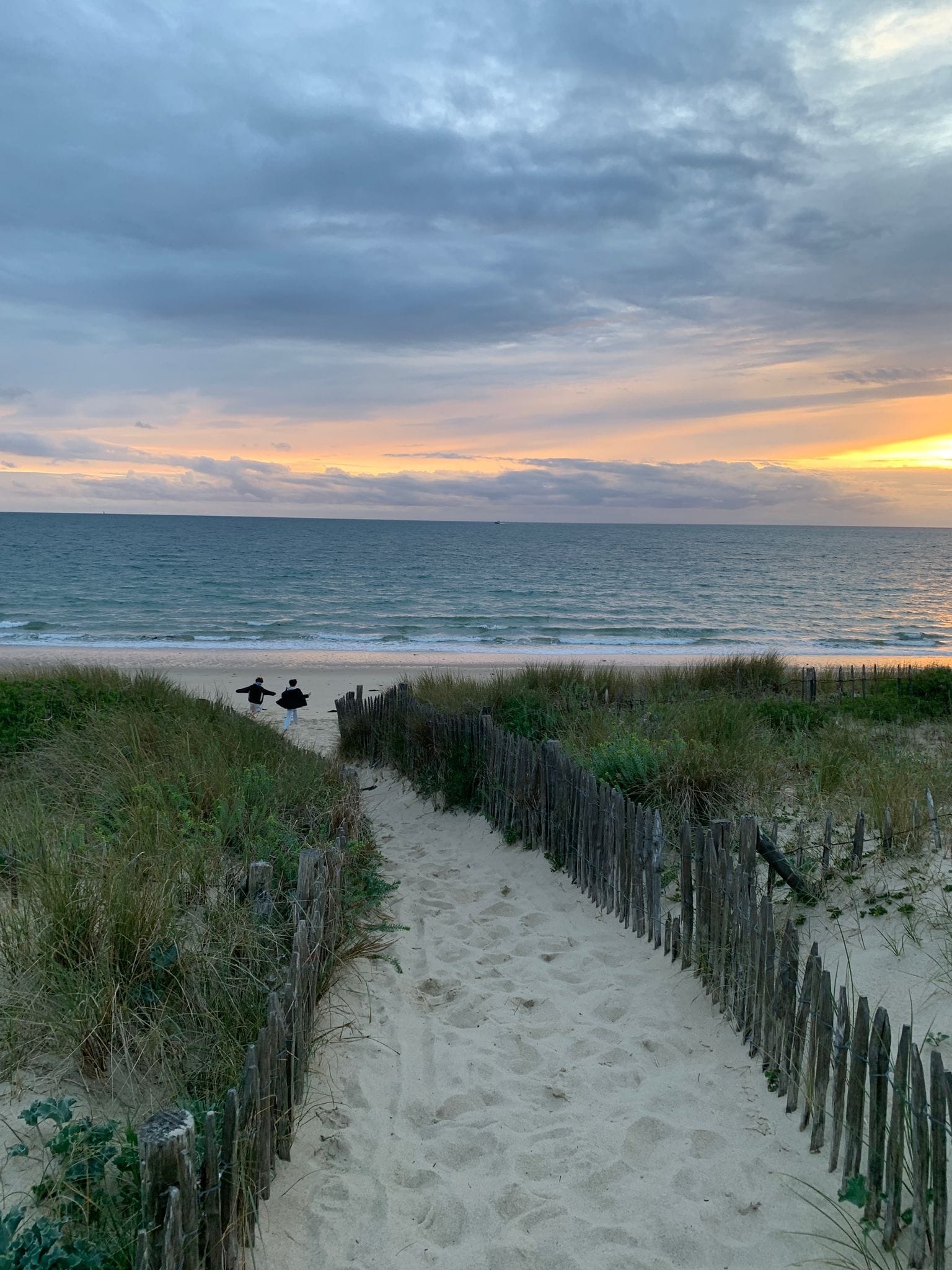 A sandy pathway leading to the beach with a wooden fence on one side.