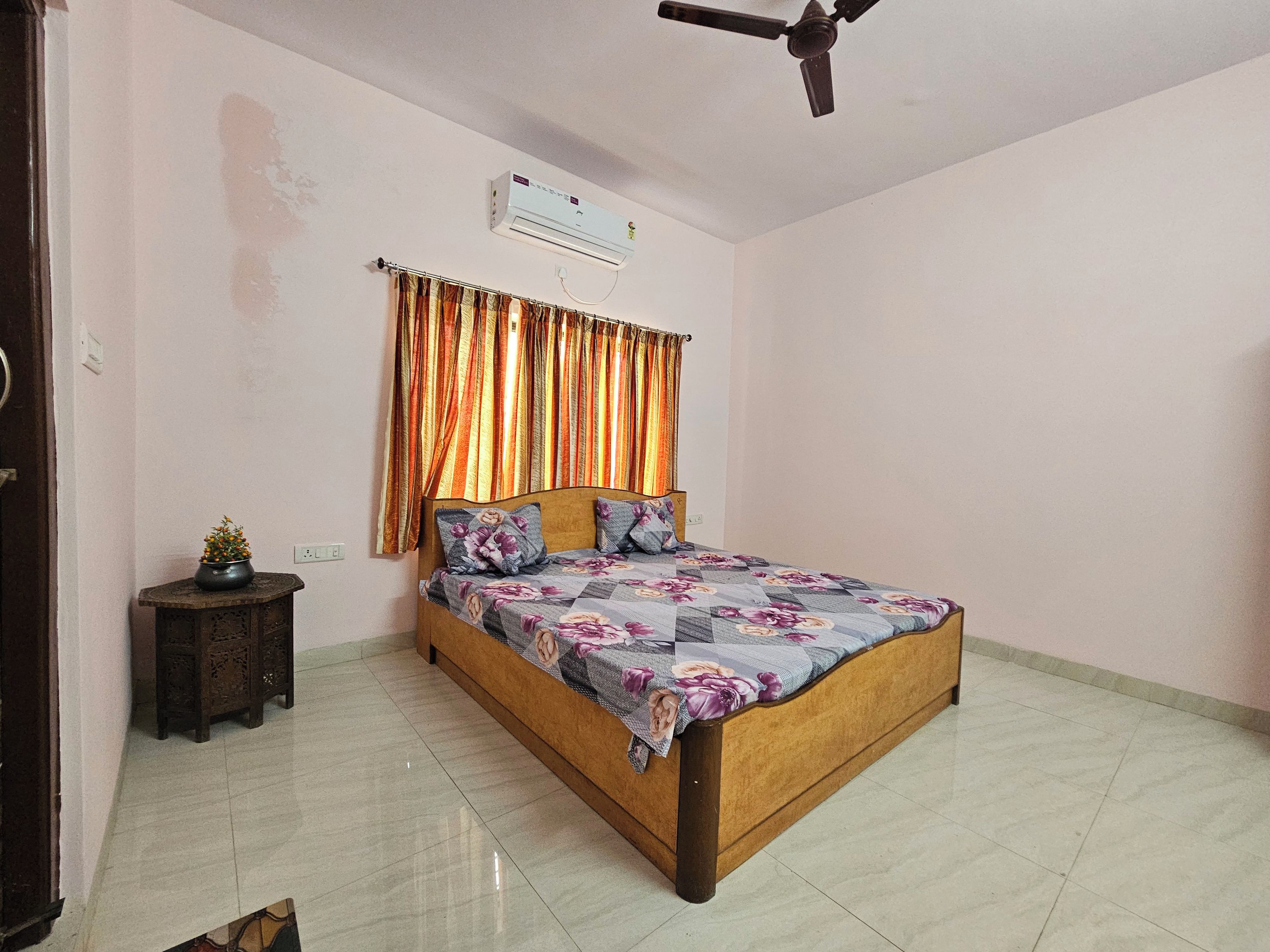 A bedroom with a bed, a ceiling fan, an air conditioner, and a small table with a plant.