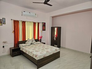 Quarto com cama, mesas de cabeceira, ventilador de teto, ar condicionado, cortinas, espelho e porta.
