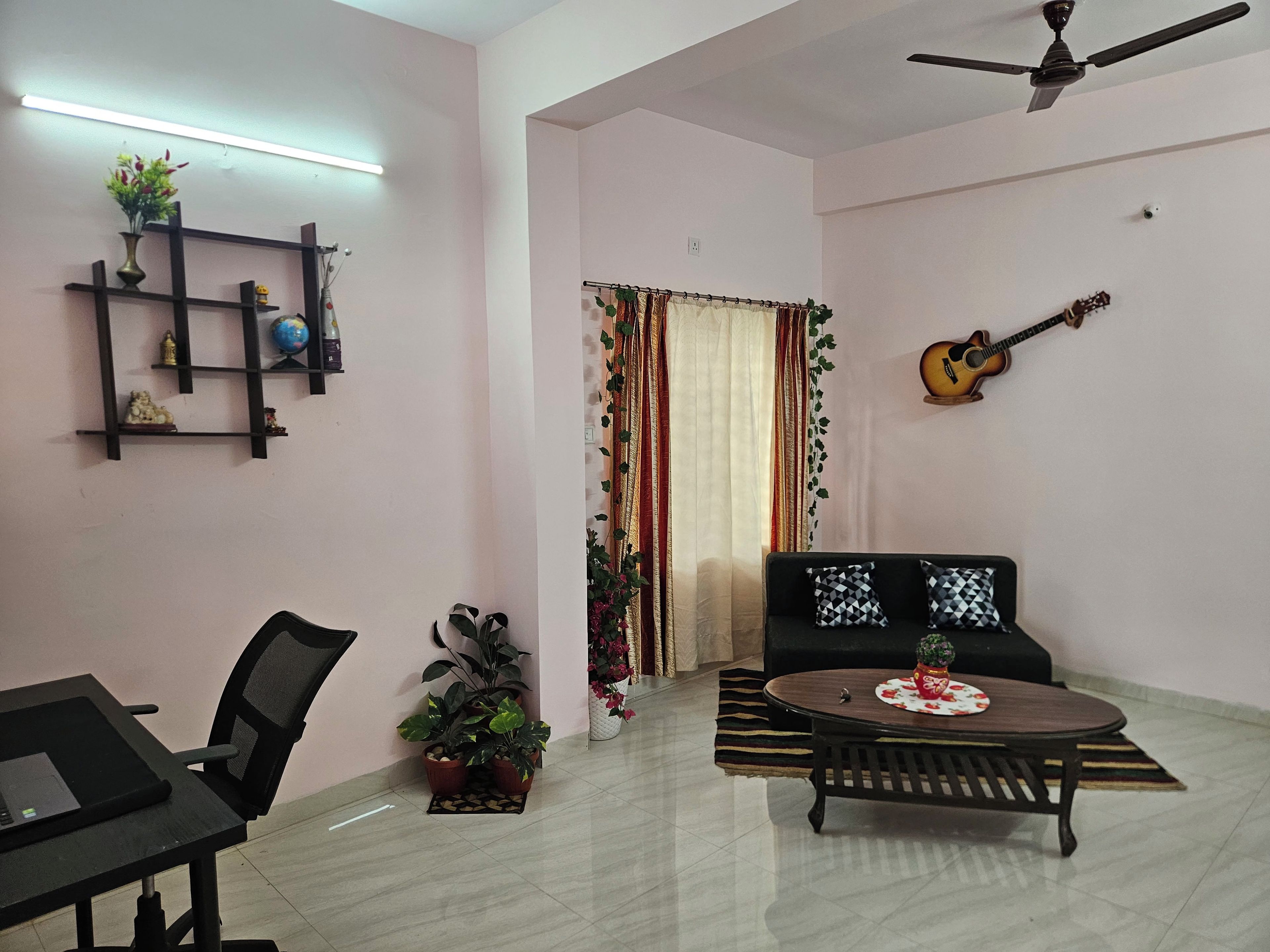 Ein Wohnzimmer mit einer Couch, einem Couchtisch, einem Laptop auf einem Schreibtisch, einer Gitarre an der Wand und einem Deckenventilator.