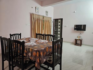 Uma área de refeições com uma mesa posta para quatro pessoas, rodeada por cadeiras, uma televisão presa na parede e uma porta com padrão listrado.