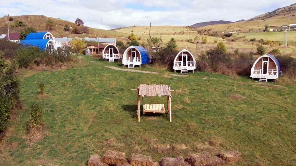 A rural setting with several small, dome-shaped cabins with white railings, a wooden bench, and hay bales in a grassy field.