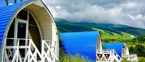 A row of rustic, blue-roofed cabins with white railings, set against a backdrop of green hills and a cloudy sky.