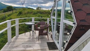 A wooden deck with a glass enclosure, wicker chairs, and a view of a green landscape with mountains.