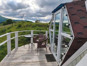 A wooden deck with a glass enclosure, wicker chairs, and a view of a green landscape with mountains.