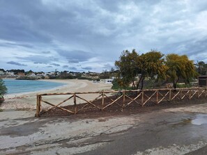 A beach with a wooden fence, a paved path, and a view of the sea and distant buildings.