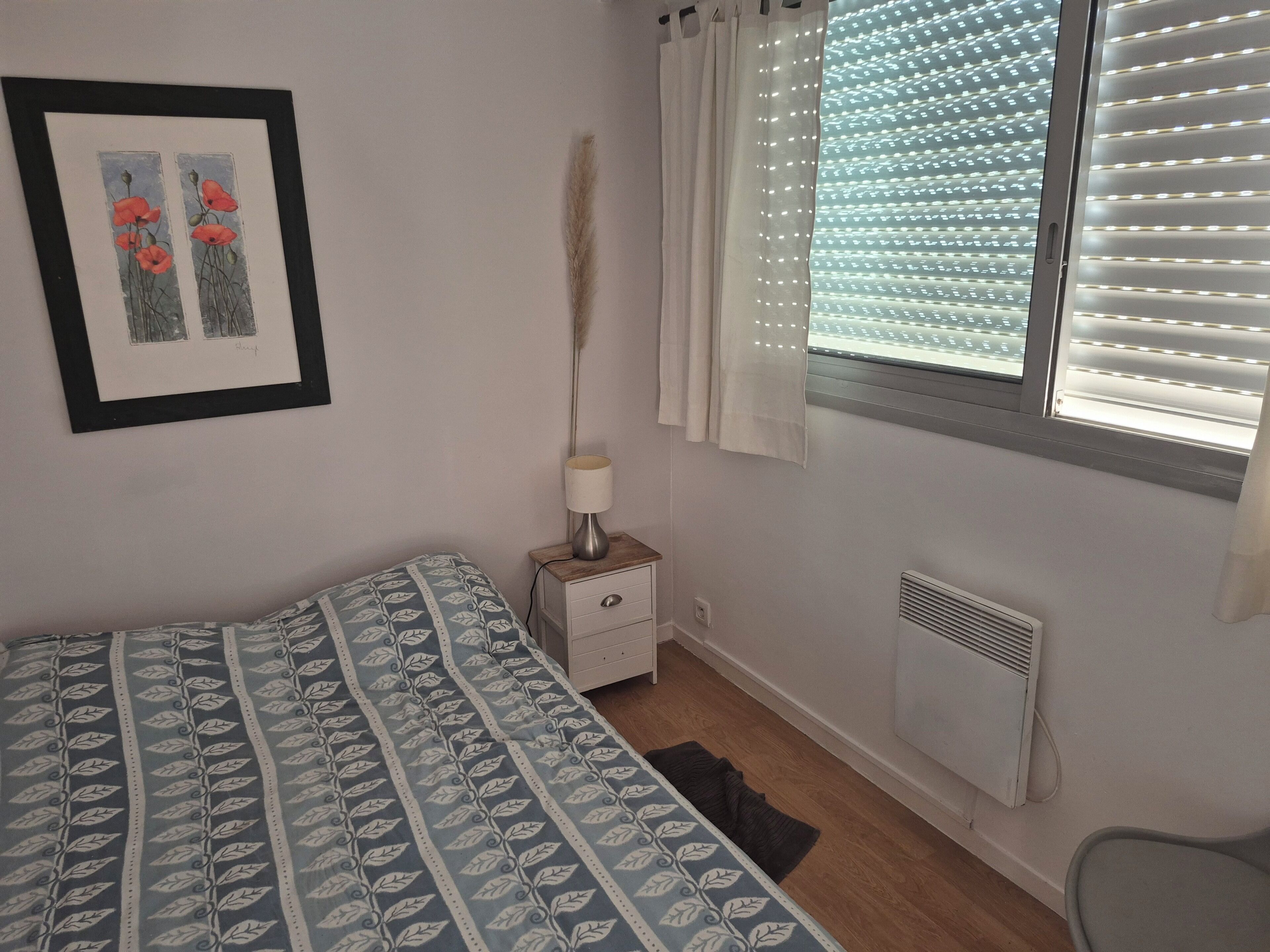 Une chambre à coucher avec un lit, une table de chevet avec une lampe, un radiateur et une fenêtre avec des stores.