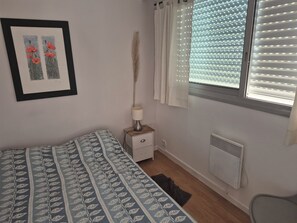 A bedroom with a bed, a bedside table with a lamp, a radiator, and a window with blinds. - Charming, very bright one-bedroom apartment (Villeneuve-Loubet)