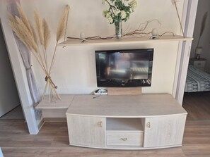 A modern living room with a flat-screen TV mounted on a wooden shelf, a light-colored console table with drawers, and decorative plants. - Charming, very bright one-bedroom apartment (Villeneuve-Loubet)