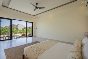 A modern bedroom with a large bed, a ceiling fan, and a view of a pool and greenery outside. - Amani Estate (Badung)