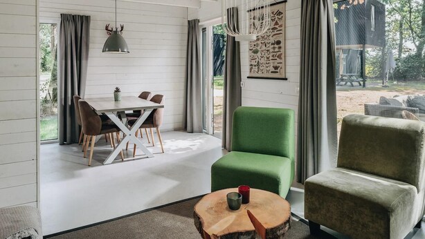 Een modern interieur met een eethoek, een groene fauteuil, een ronde houten tafel en een groot raam met gordijnen.