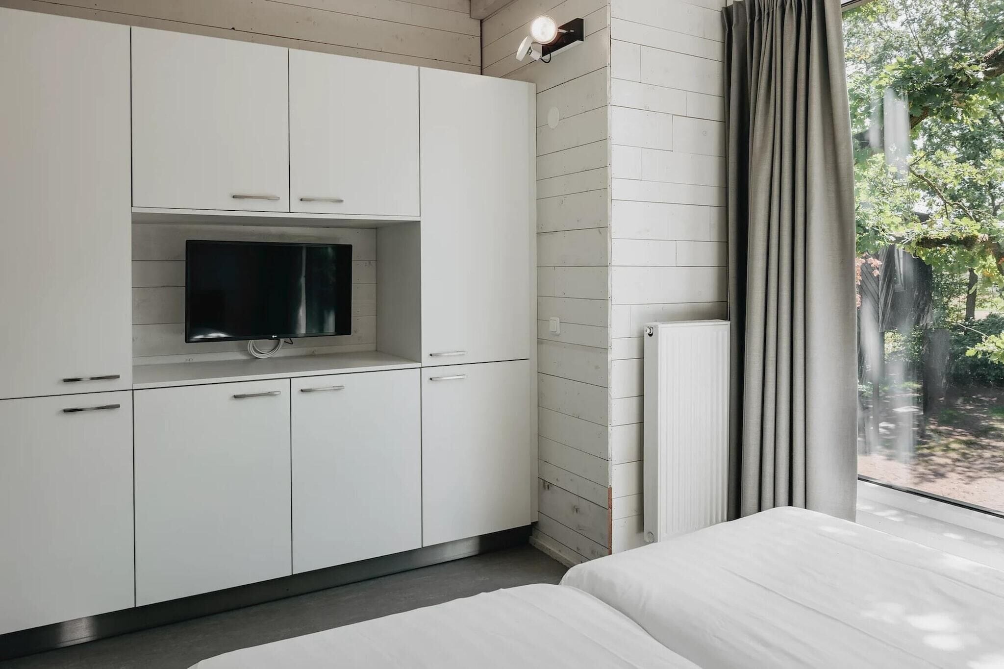 Ein Schlafzimmer mit einem Bett, einem an einer Kommode befestigten Fernseher und einem Fenster mit Vorhängen.